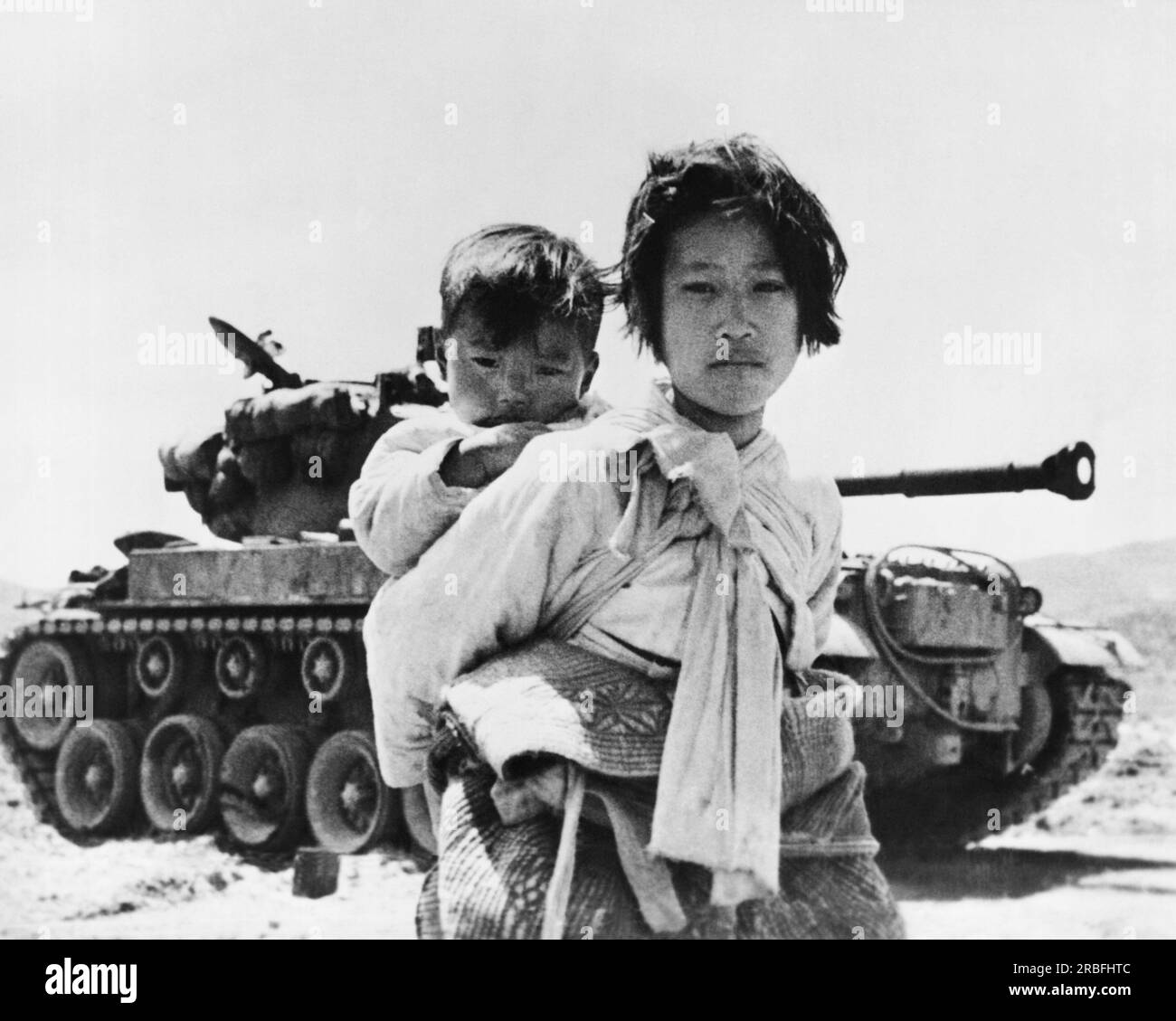 Haengju, Korea:  June 19, 1953 A nine year old Korean refugee girl carrying her small brother trudges past a Patton tank. The rest of her family has been killed in the war. Stock Photo