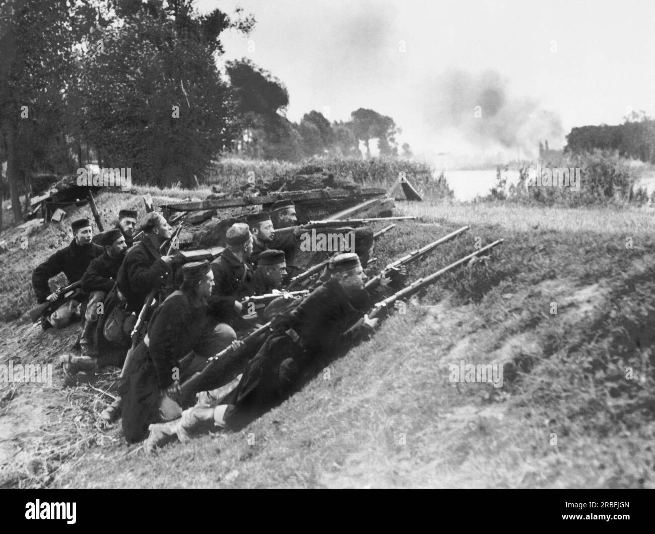 Belgium:  October 16, 1914 Belgian marksmen with their long bore rifles lay in ambush on the bank of the Nethe ready to pick off the Germans on the opposite side. The smoke in the background is from buildings that the Germans have set on fire. Stock Photo