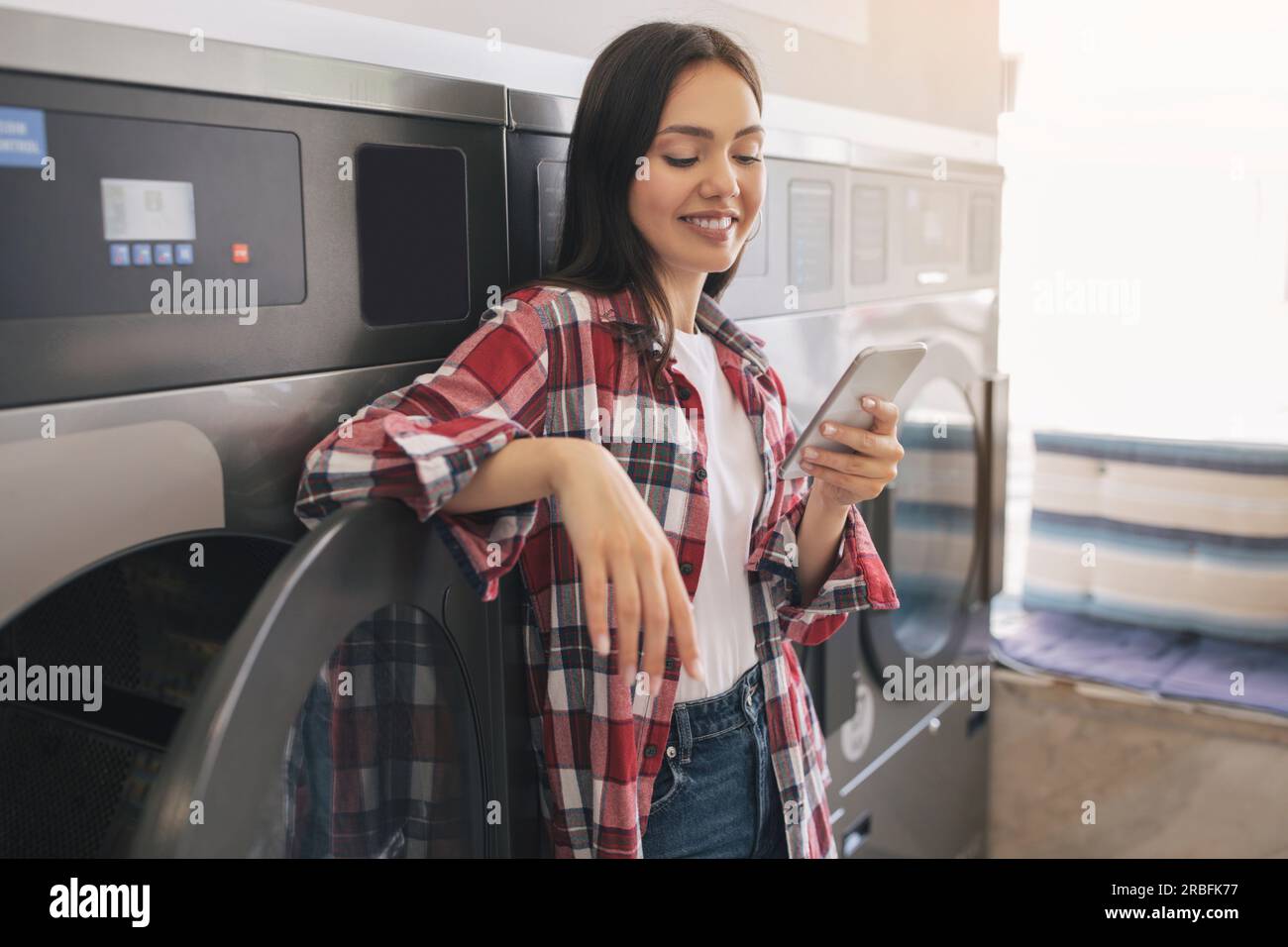 Happy Young Lady Using Cellphone While Doing Laundry Indoor Stock Photo