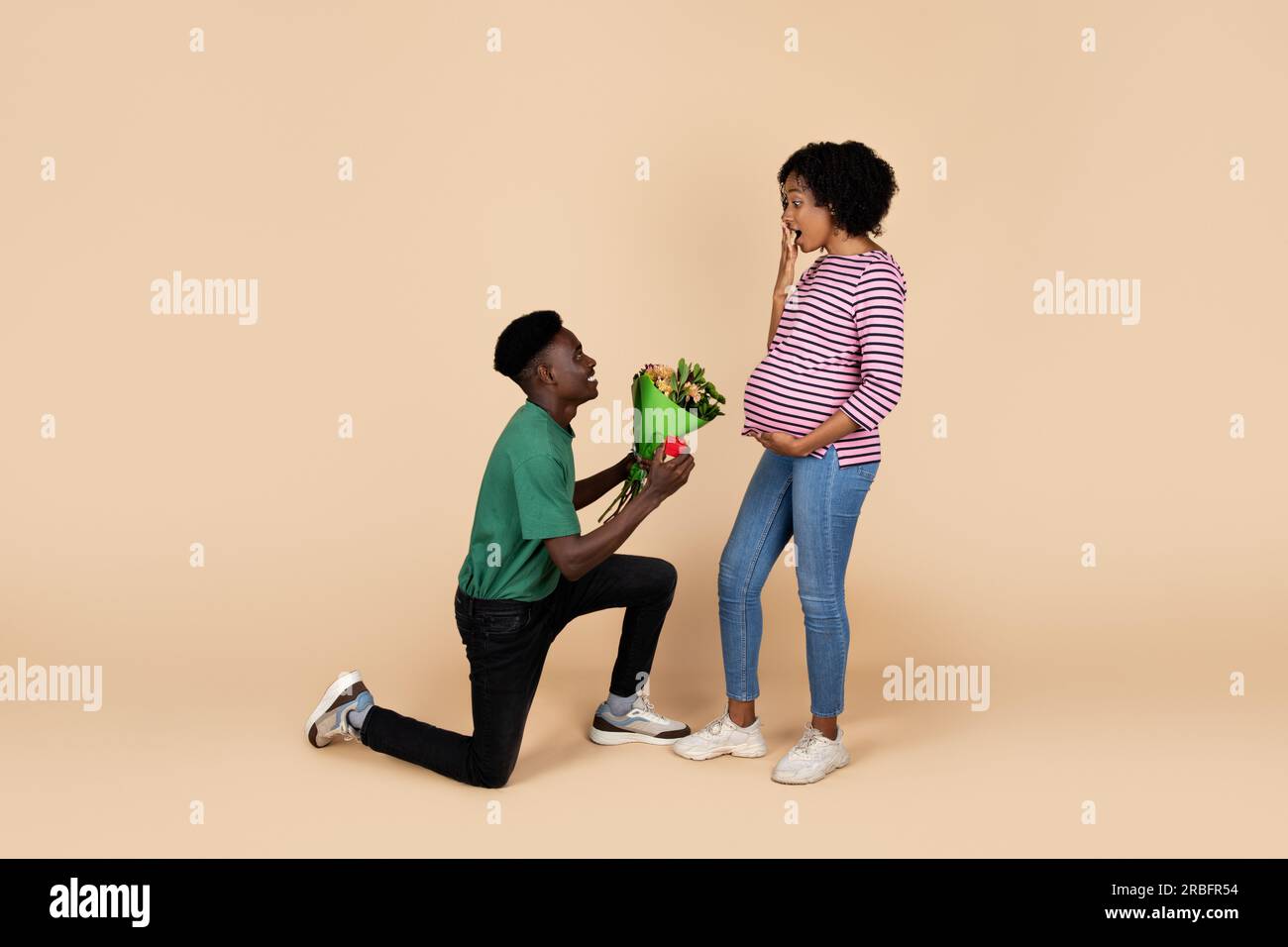 Happy millennial african american husband kneeling gives bouquet of flowers to woman with big belly Stock Photo