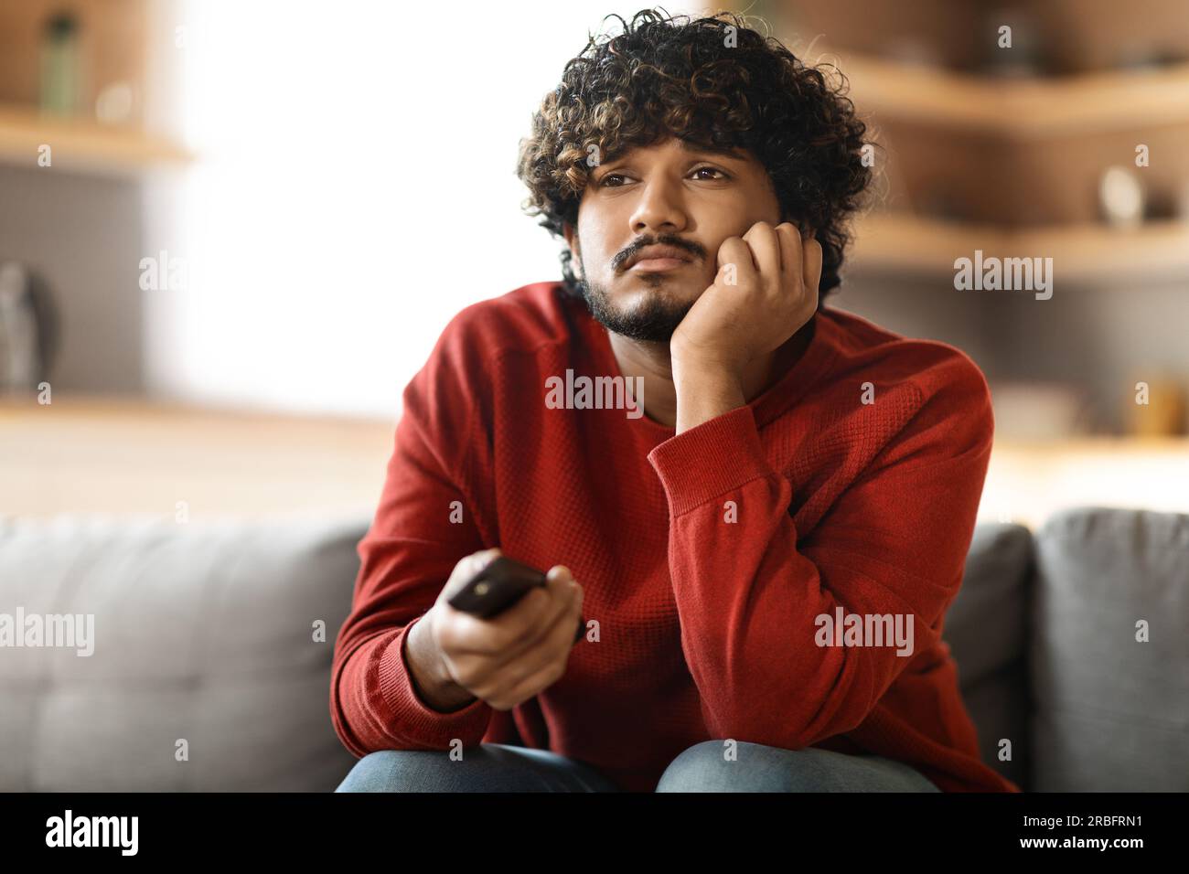 Portrait of bored indian man watching TV at home, sitting on sofa Stock Photo