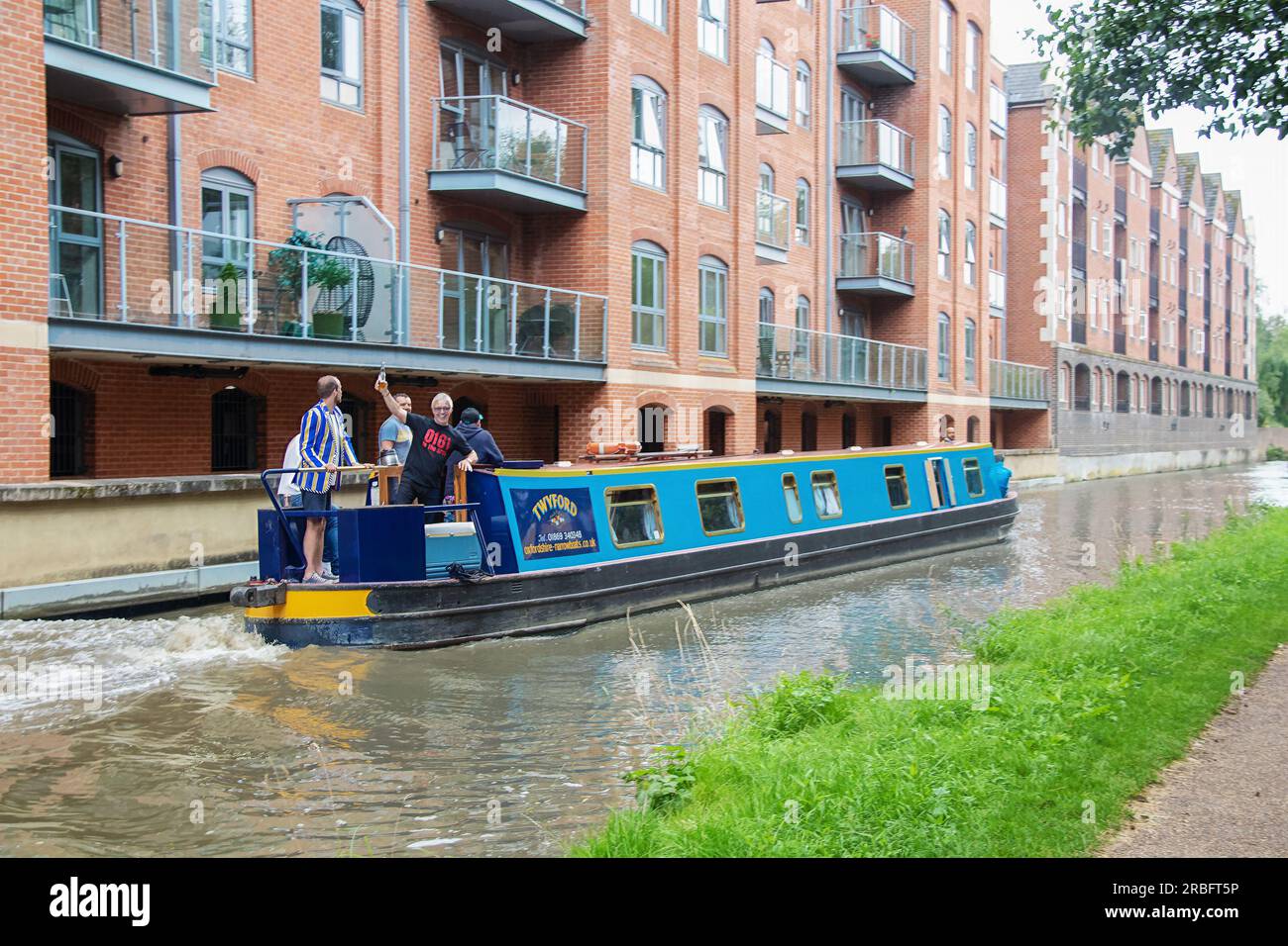 7-27-2019 Oxford England -Men partying on river boat on river passing overlooking apartments with one man wearing school jacket and shorts and another Stock Photo