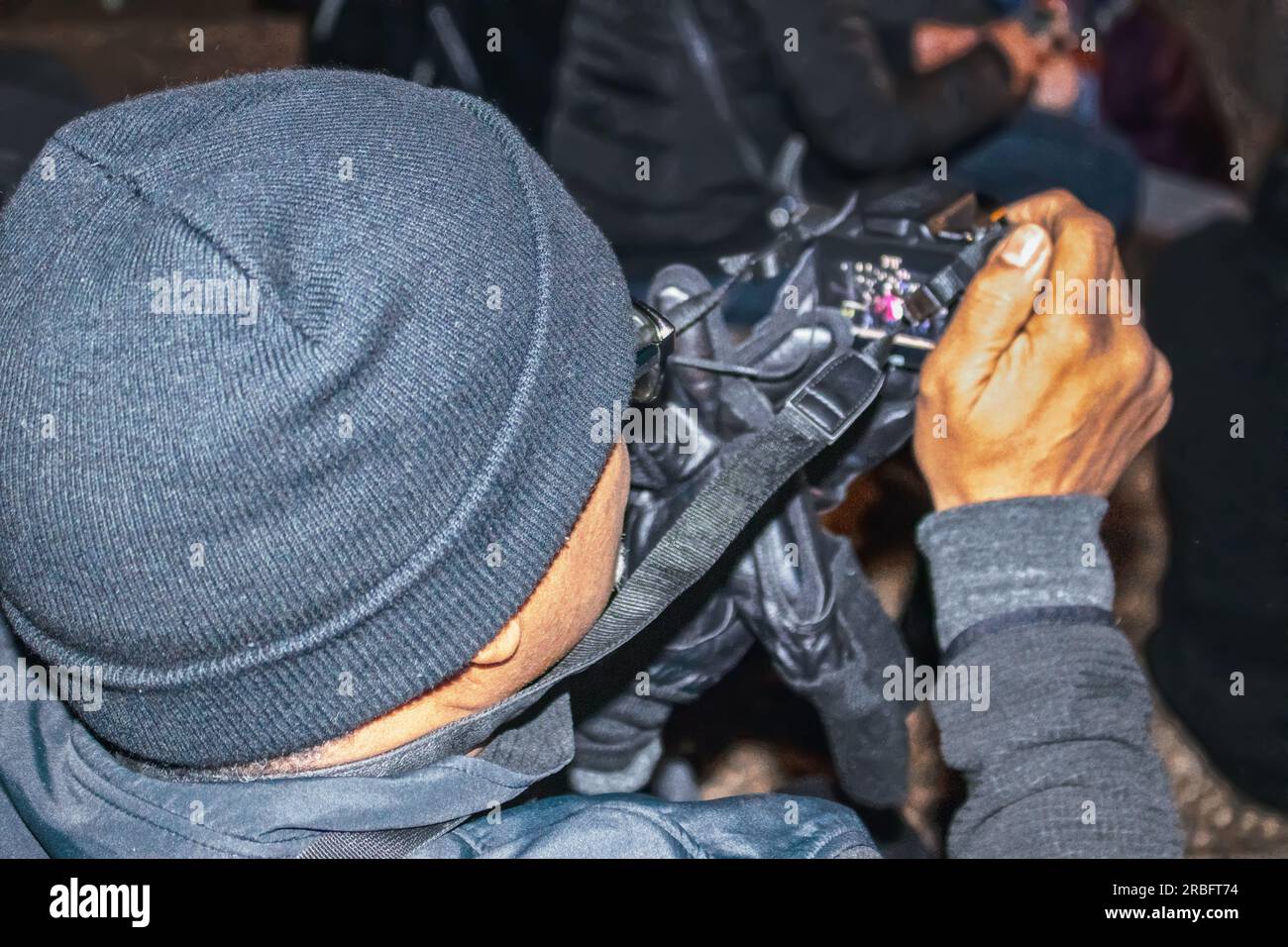 Black man with knitted cap videotaping a performance at night - shot from over his shoulder featuring camera Stock Photo