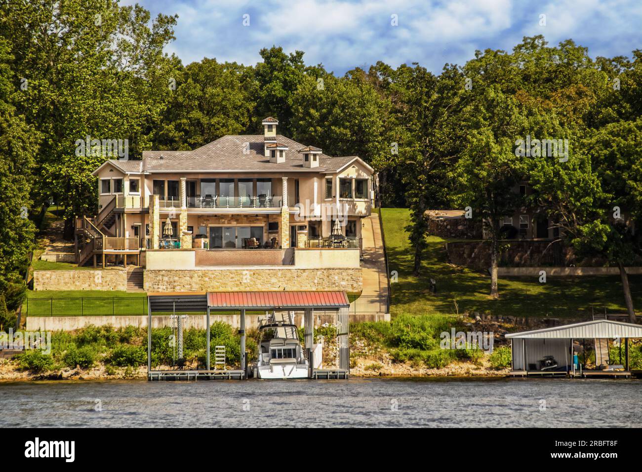 Huge lake house with two layers of deck toward water with big motor yacht parked in private dock and beautiful trees in background Stock Photo