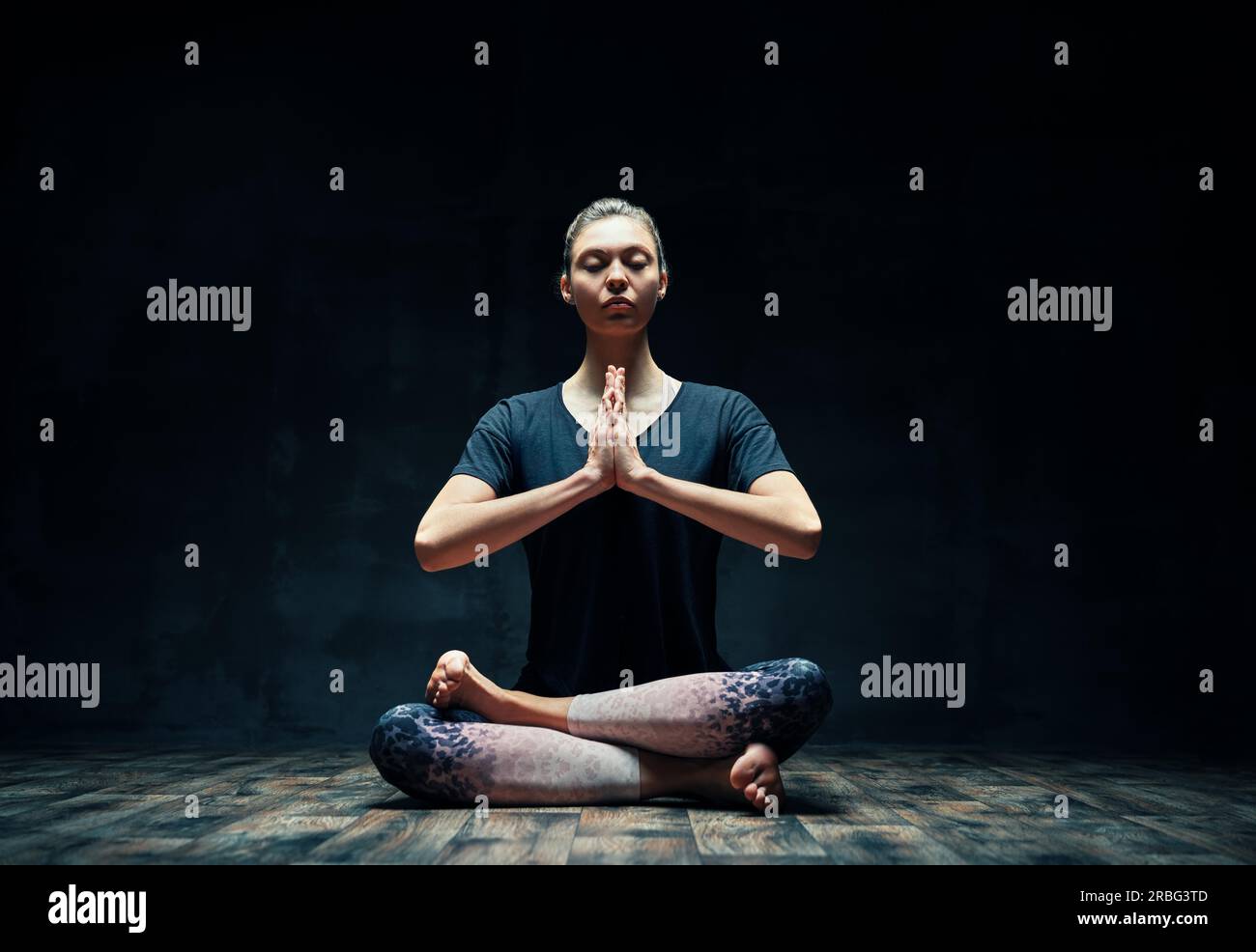 Young sporty woman practicing yoga doing Ardha Padmasana exercise with namaste in dark room. Lotus pose, meditation concept Stock Photo