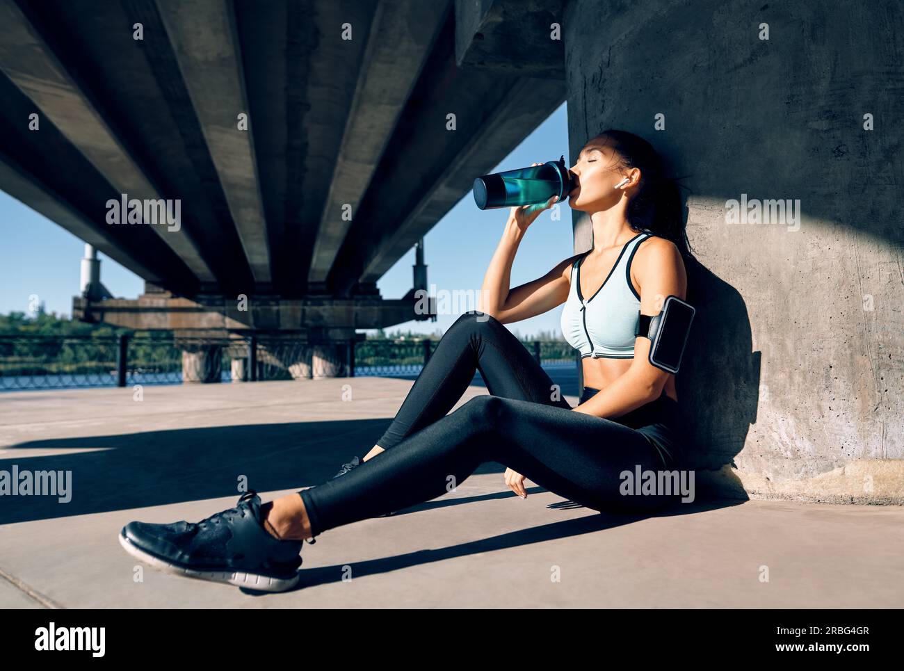 Young fitness woman drinking water from bottle. Sporty female resting after work out exercising in industrial city background near river Stock Photo