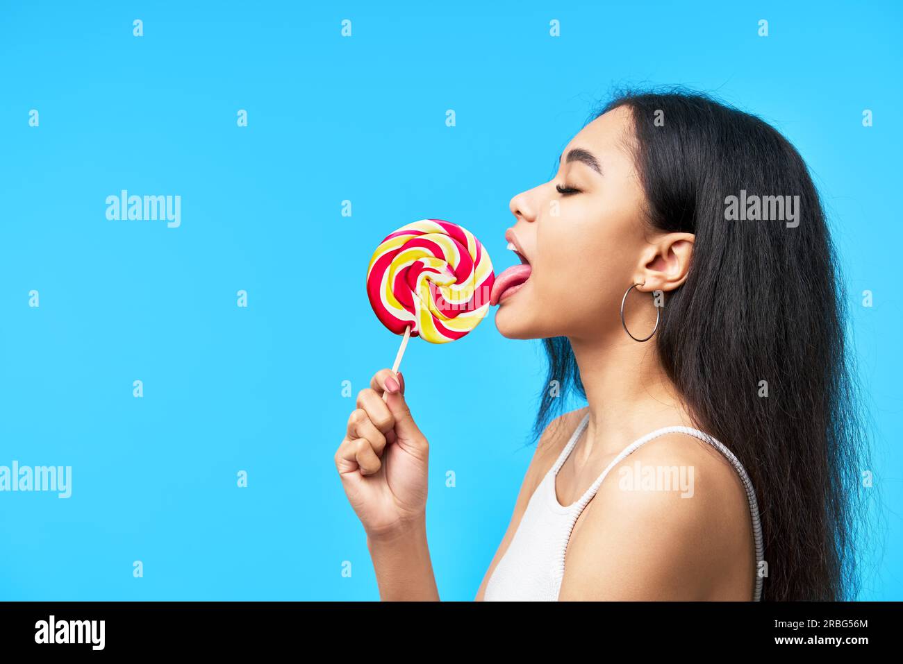 Profile view of young black woman licking colorful lollipop on blue background with copy space. Food, fun concept Stock Photo