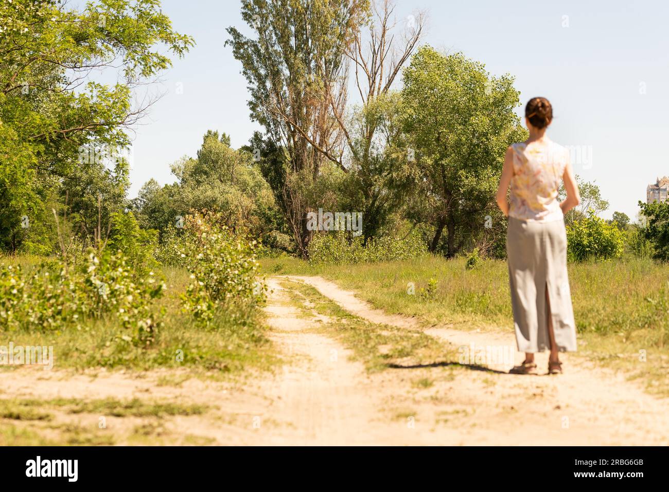 A woman with a chignon, standing up close to the country road in Kiev, Ukraine, observes the trees in the distance. The silhouette of the lady is out Stock Photo