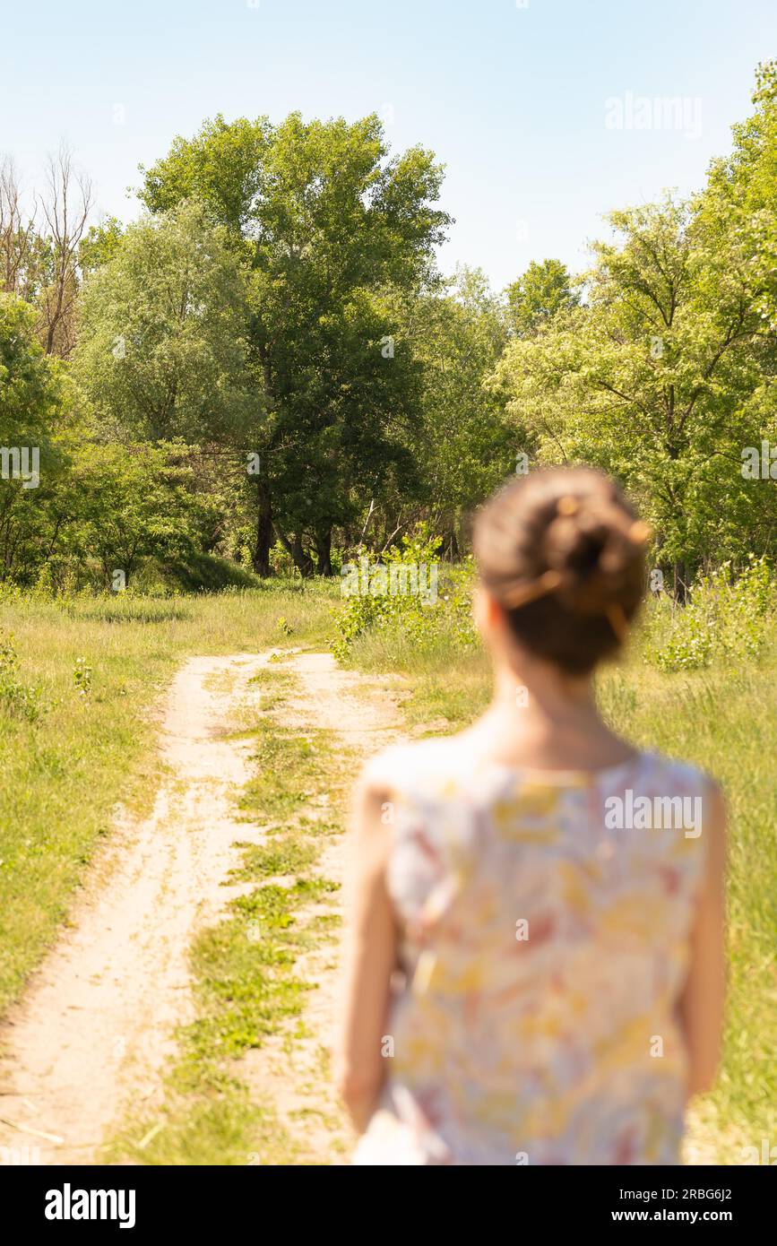 A woman with a chignon, standing up close to the country road in Kiev, Ukraine, observes the trees in the distance. The silhouette of the lady is out Stock Photo