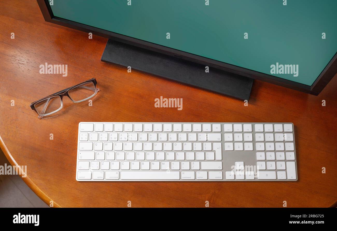 A computer monitor, a digital wireless keyboard and eyeglasses on a round wooden desk Stock Photo