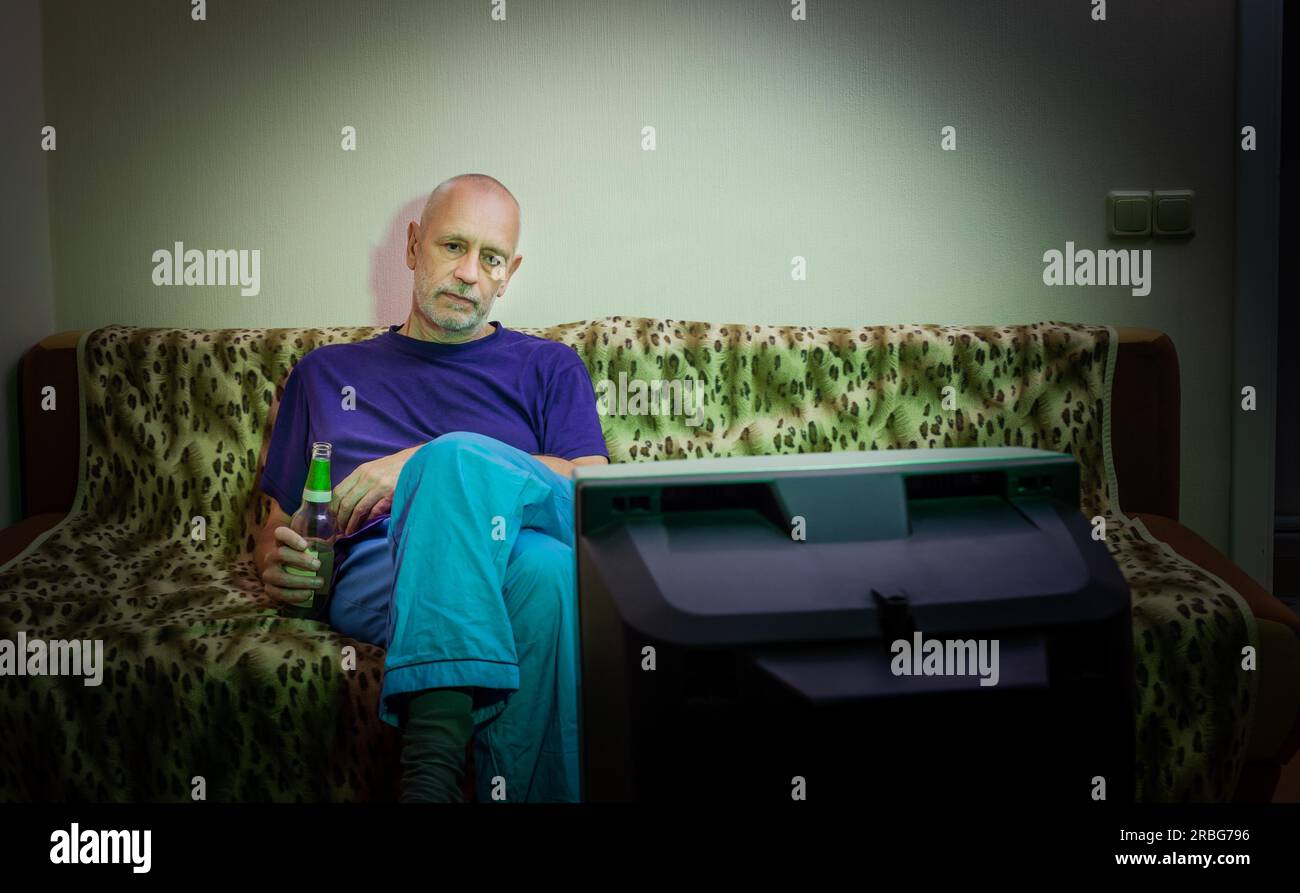 An adult man, alone, watches television, looking sad, sitting on a couch, with a bottle of beer in the hand. He watches a sad movie or listens to bad Stock Photo