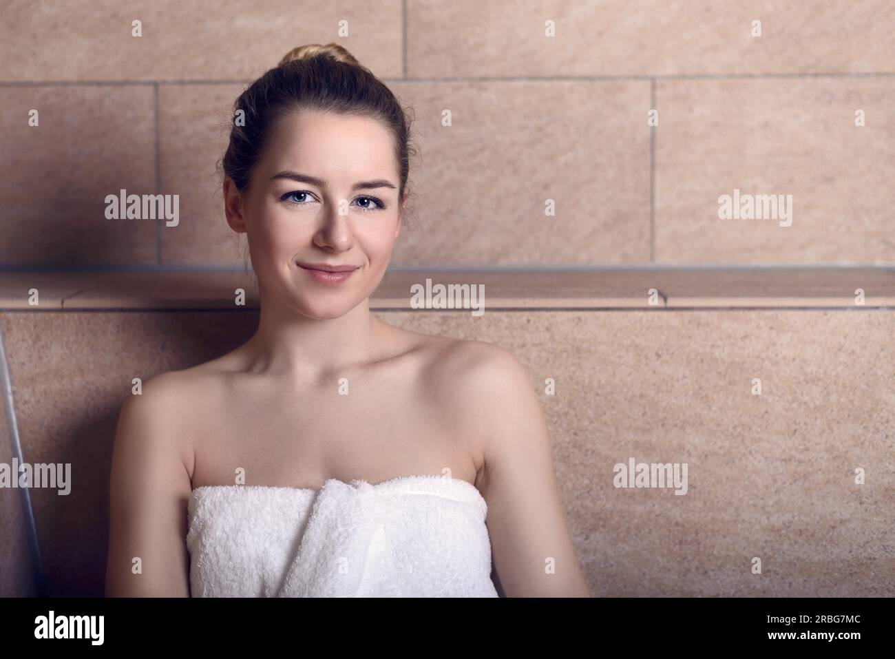 Attractive smiling young woman in a bath towel wrapped around her chest standing in a light brown tiled bathroom in a health and beauty concept Stock Photo