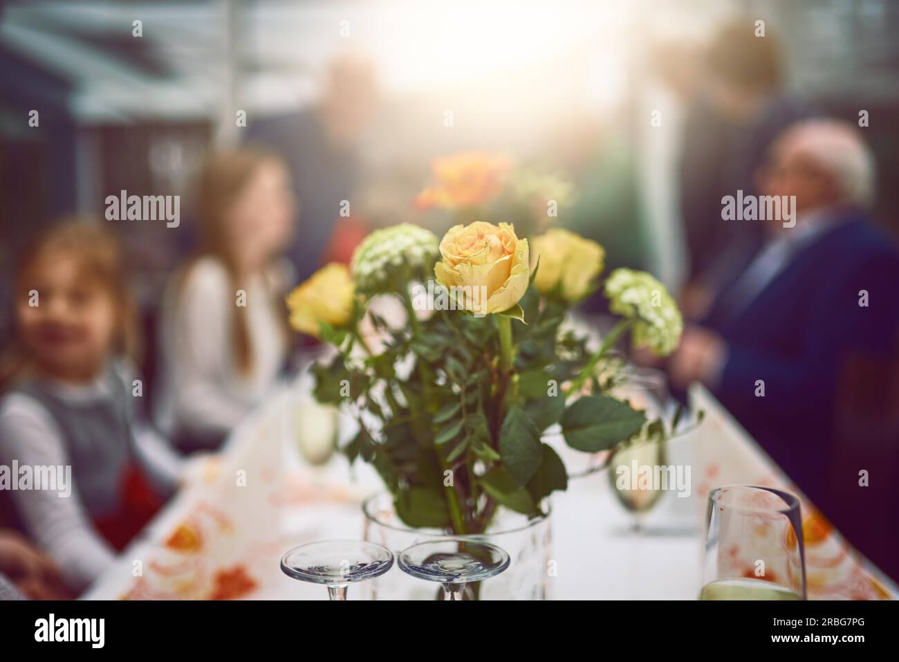 Vase of fresh yellow and orange flowers as a table centerpiece at a dinner party with selective focus to the arrangement and blurred people Stock Photo