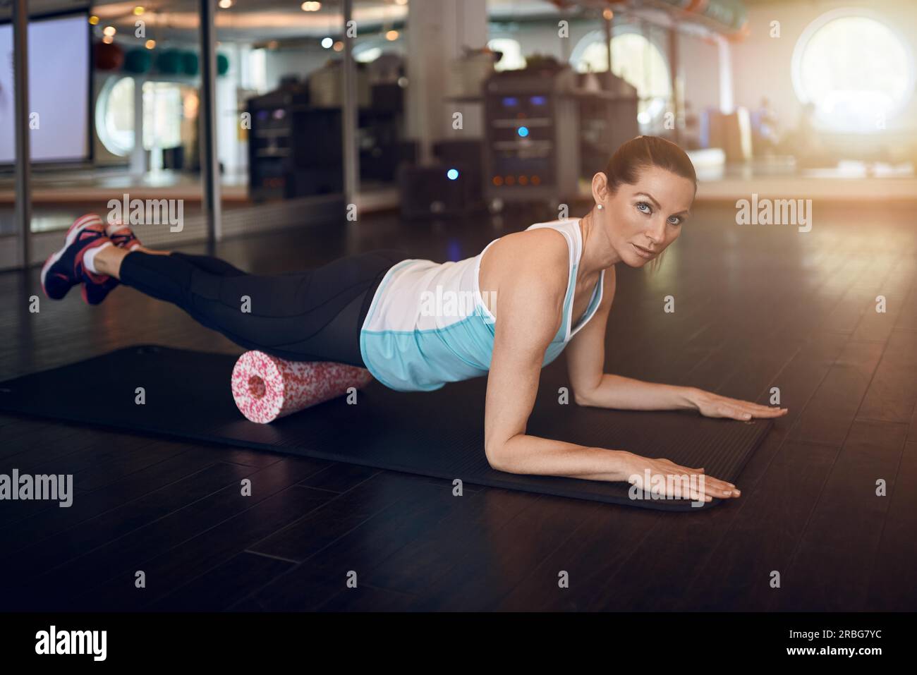 Woman in mint sleeveless shirt and black leggings in gym doing exercise with foam or fascia roll and looking at camera Stock Photo