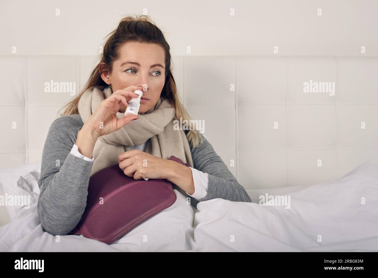 Sick woman wrapped in a warm scarf and clutching a hot water bottle sitting using nasal spray with a miserable expression in a seasonal healthcare or Stock Photo