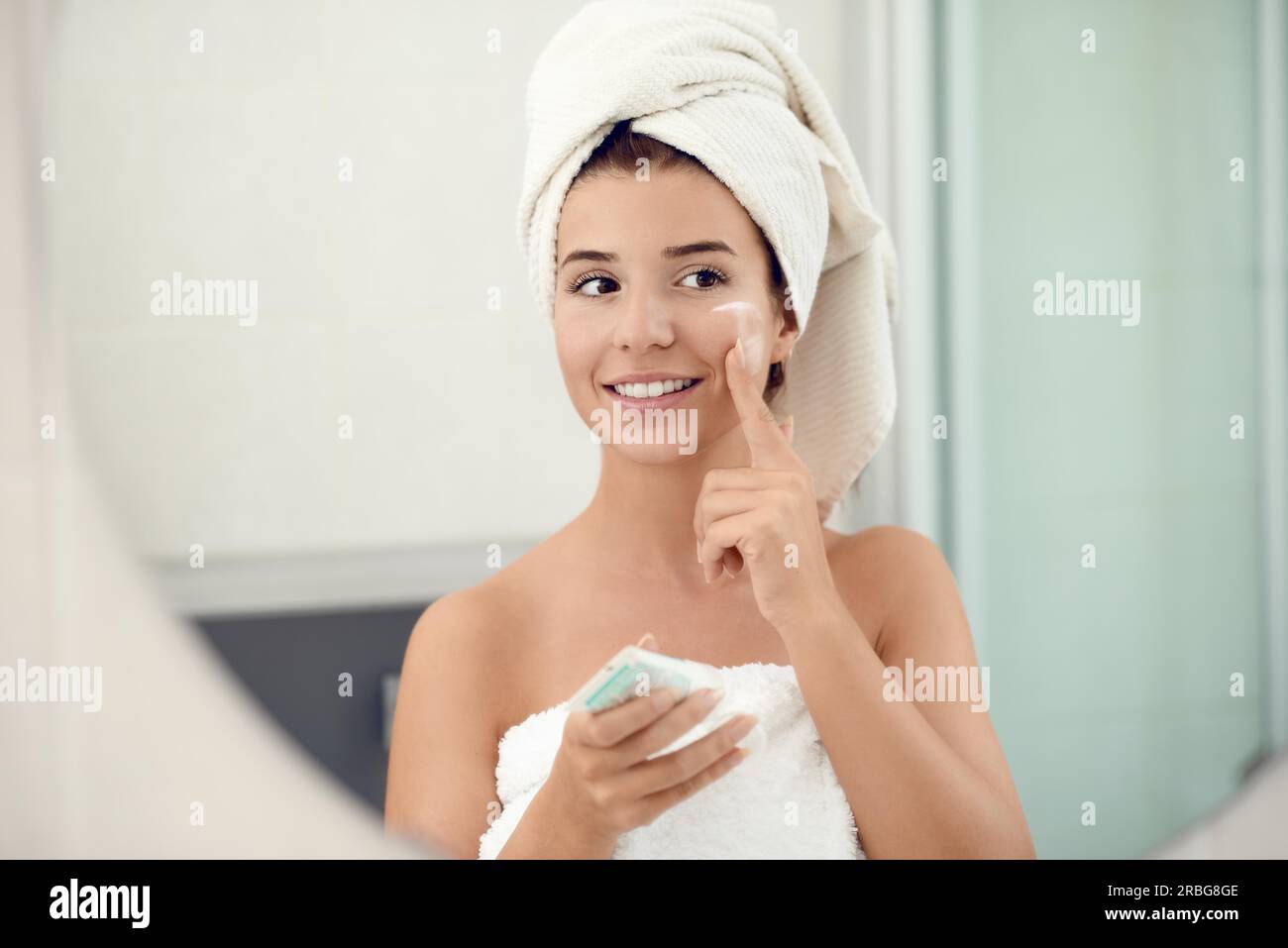 Pretty young woman in white towels in a bathroom giving the camera a lovely friendly smile as she applies cream to her cheeks while bathing in a skin Stock Photo