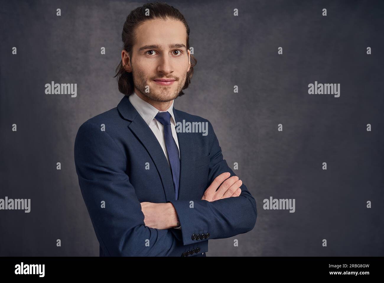 Handsome young man with long haircut in business suit with blue necktie, standing with his arms crossed against grey background with copy space and Stock Photo