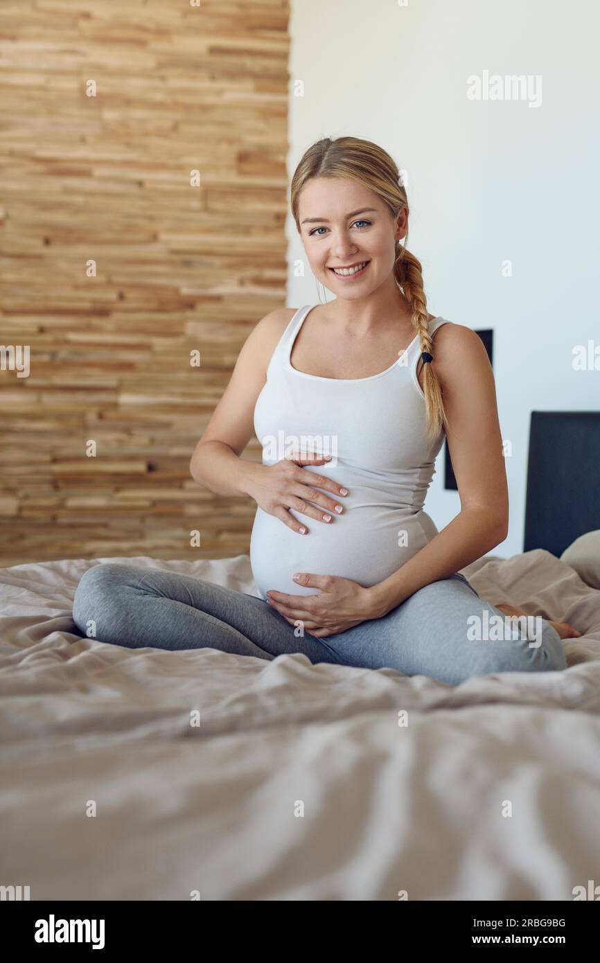 Happy pregnant woman sitting on a bed cradling her baby bump with her hands smiling at the camera Stock Photo