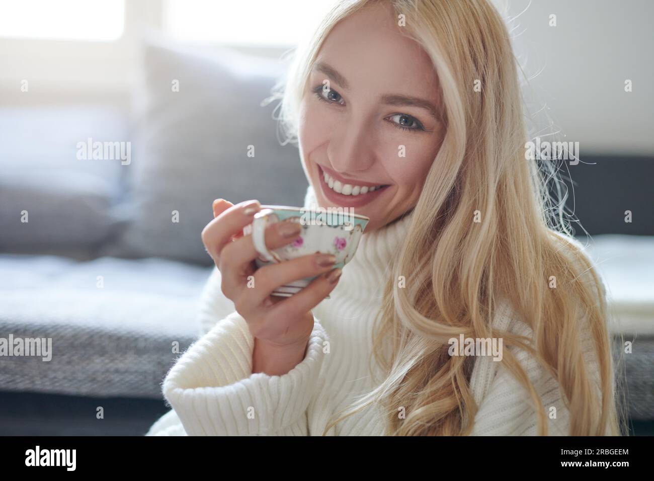 Pretty woman with long blond hair drinking tea from a porcelain cup looking at the camera with a sweet beaming smile of pleasure indoors at home Stock Photo