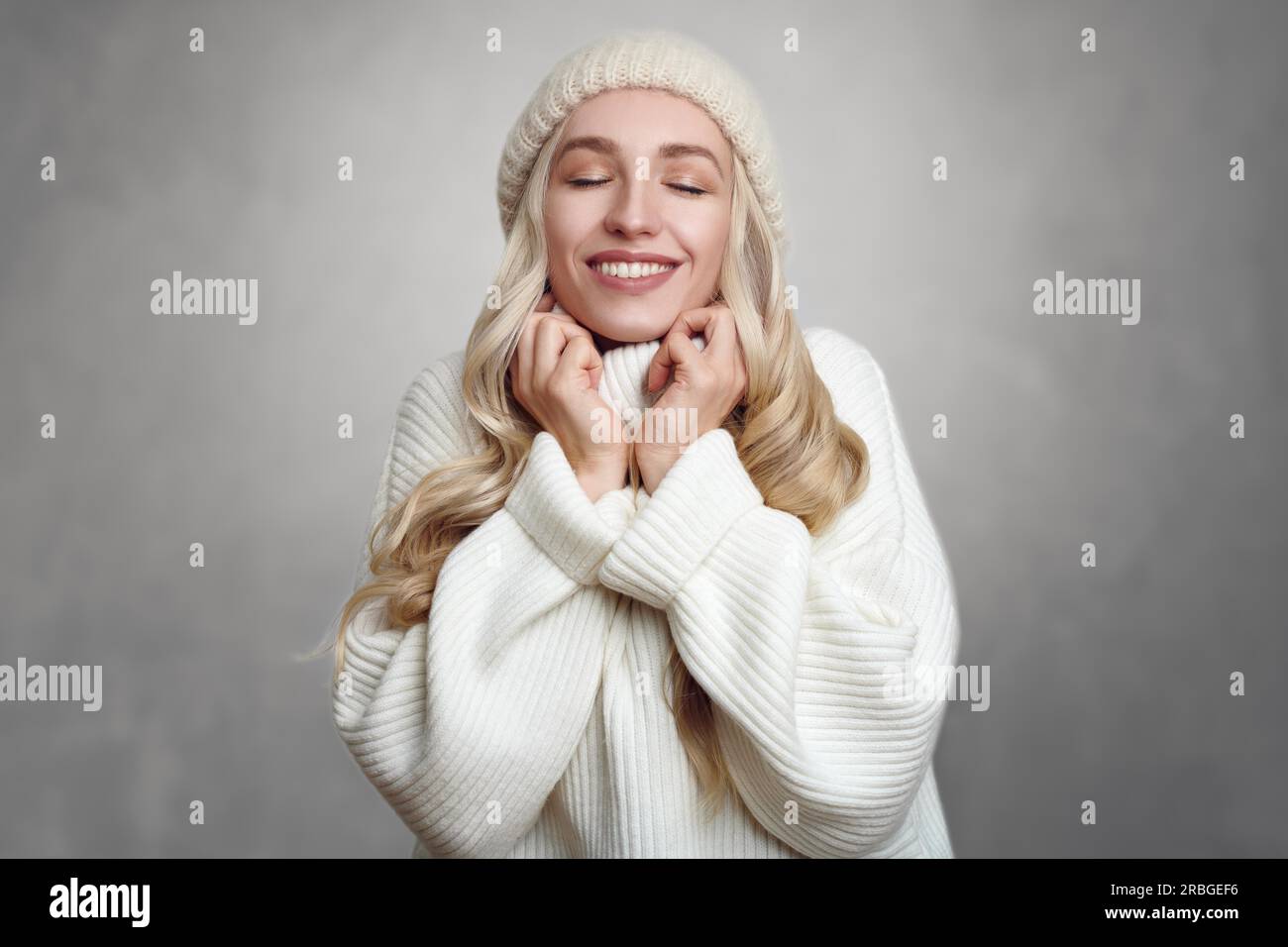 Young blond woman enjoying her white knitted sweater, standing against grey background, with her hands to her neck and smiling with eyes closed Stock Photo