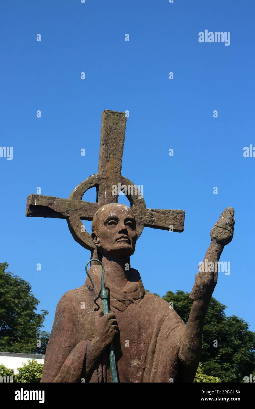 Part of statue Saint Aidan in grounds Lindisfarne Priory by artist Kathleen Parbury. Red concrete statue St. Aidan with torch, bronze crook, cross. Stock Photo