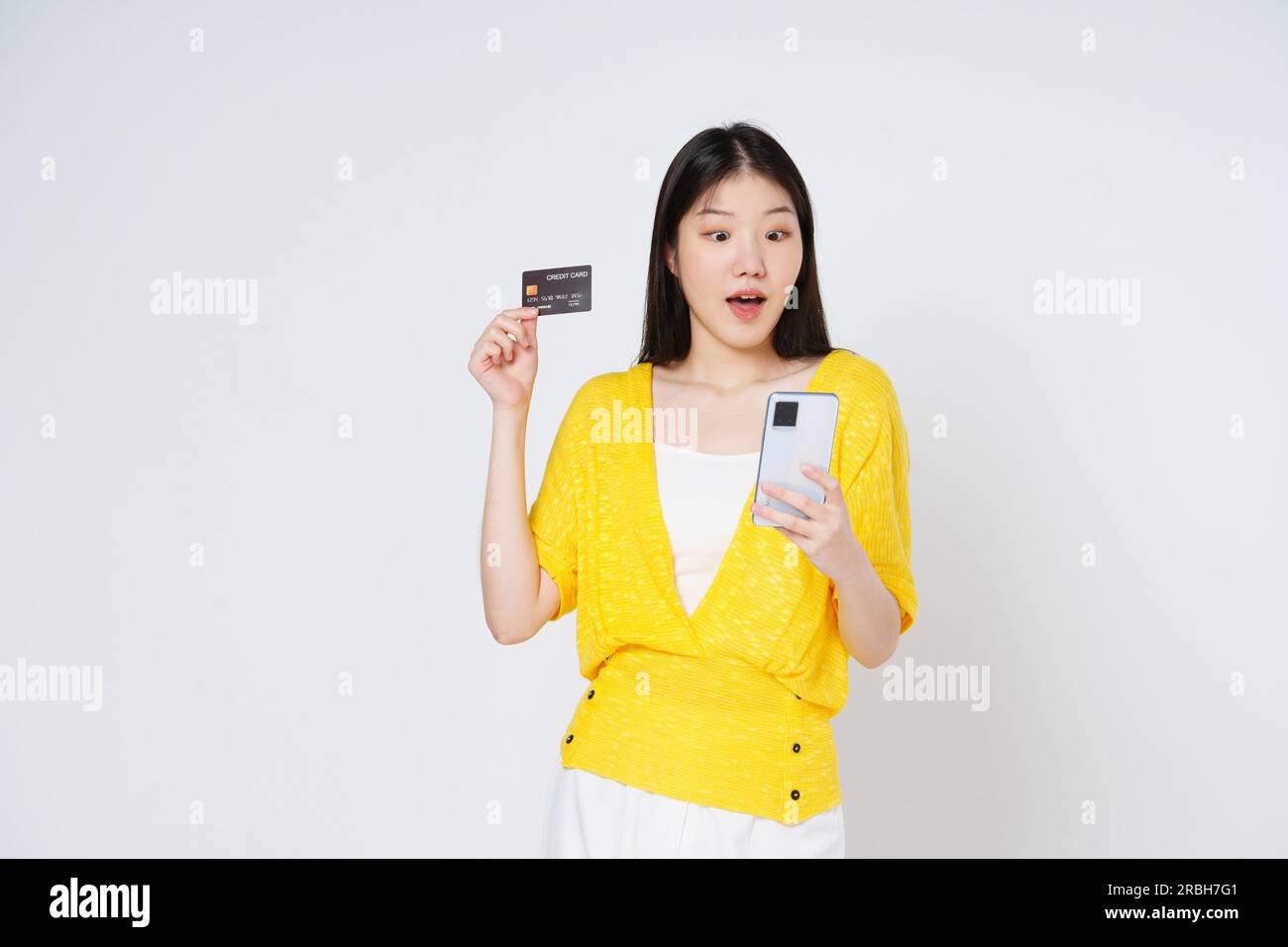 Happy young woman showing credit card while holding mobile phone isolated white background. Stock Photo