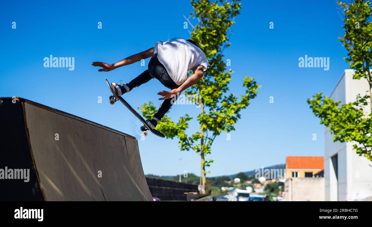 skinny skater jumping high and doing a risky trick on a blue sky with some trees Stock Photo