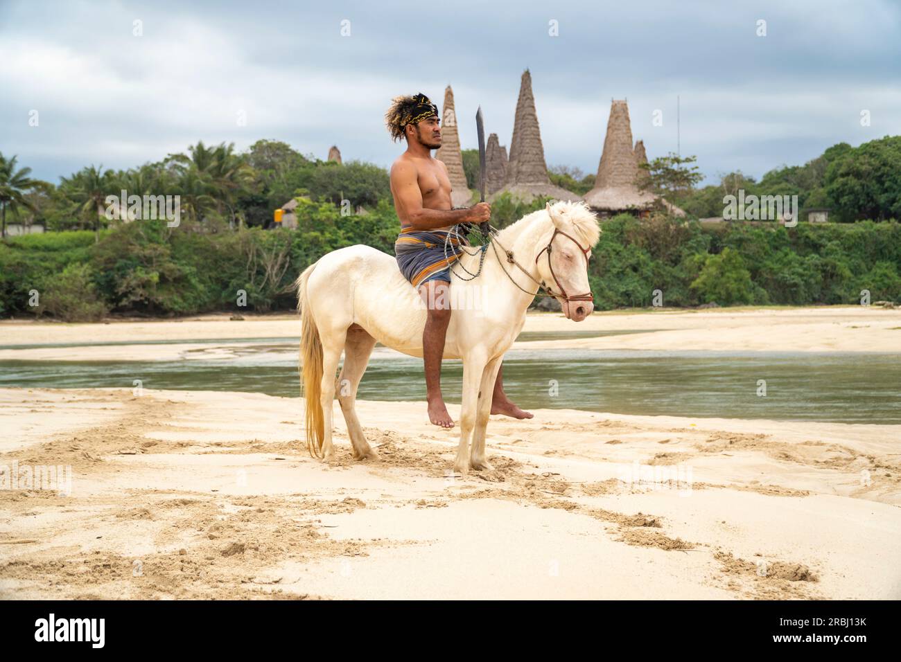 A member of the Marapu Sumba Tribe gracefully rides a horse along the shoreline, with the captivating Ratenggaro Traditional Hut in the backdrop Stock Photo