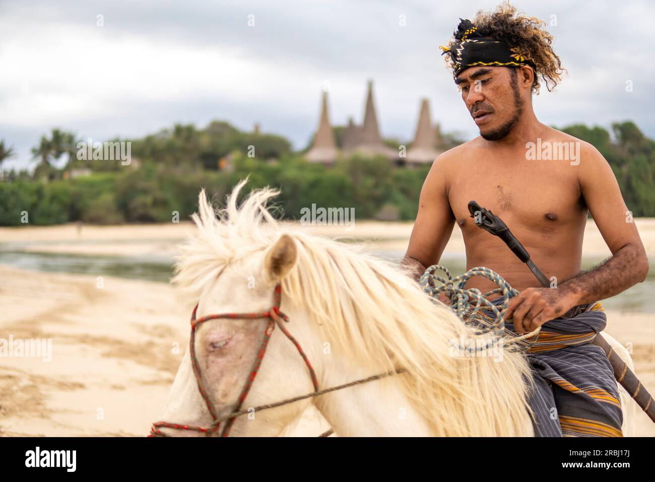 A member of the Marapu Sumba Tribe gracefully rides a horse along the shoreline, with the captivating Ratenggaro Traditional Hut in the backdrop Stock Photo