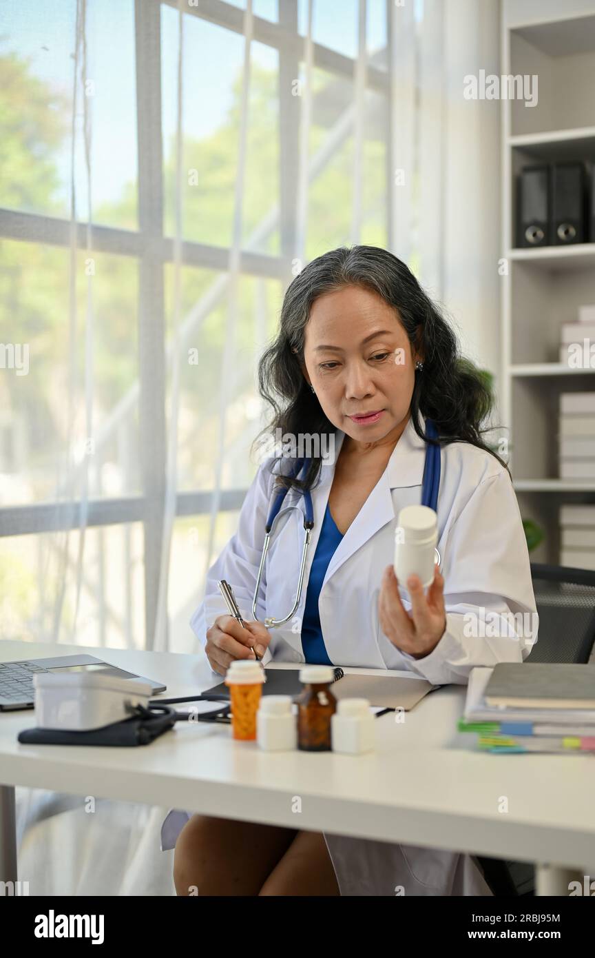 A portrait of a professional Asian senior female doctor writing medicine prescriptions and working at her desk in the hospital office. Stock Photo