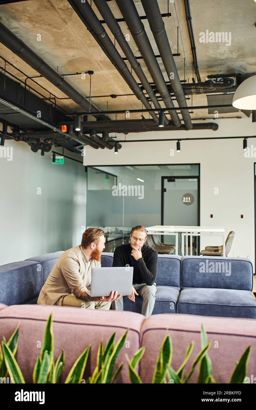 bearded businessman showing startup project on laptop to thoughtful colleague while talking on comfortable couch in lounge of modern office with high Stock Photo
