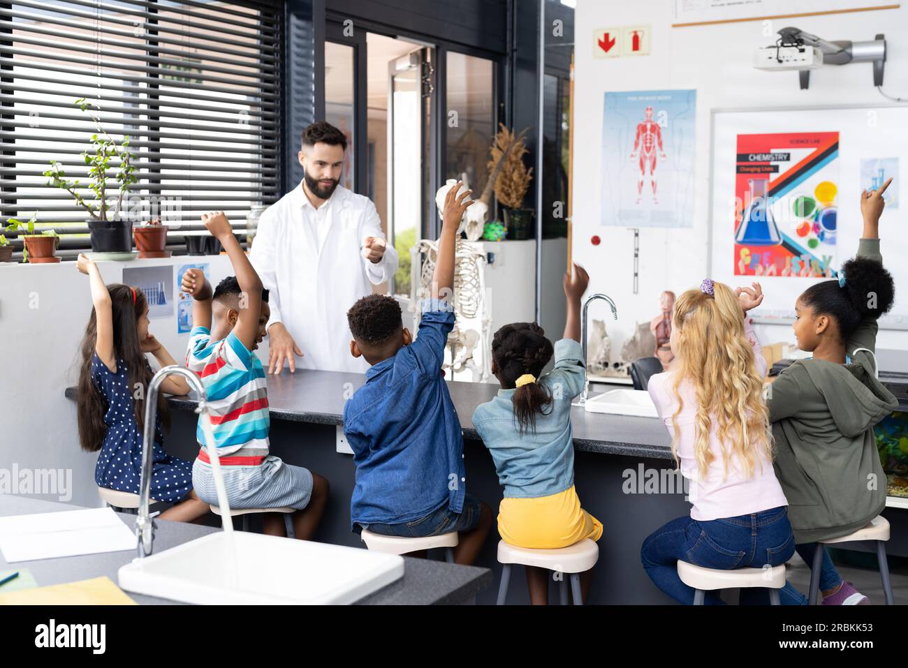 Diverse male teacher and elementary schoolchildren raising hands in ...
