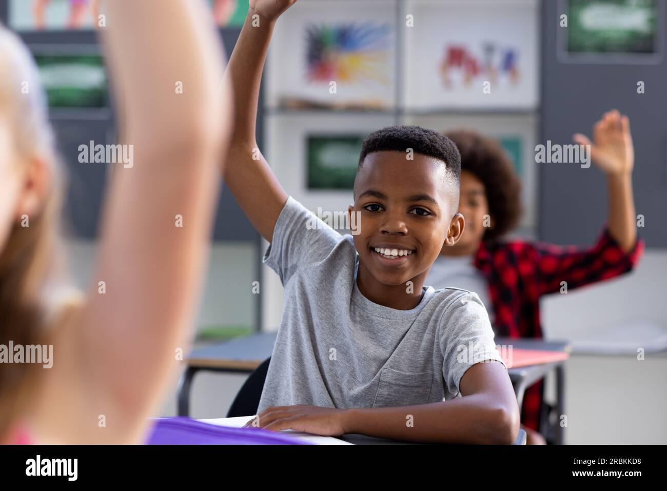 Portrait of smiling african american schoolboy sitting at desk raising ...