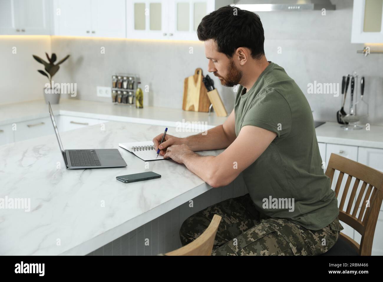 Soldier taking notes while working with laptop at white marble table in ...