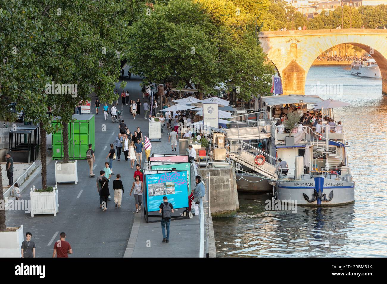 People promenade in the evening light on River Seine Right Bank on Voie Georges Pompidou with boat bars, restaurants opposite Ile Saint-Louis. Paris. Stock Photo