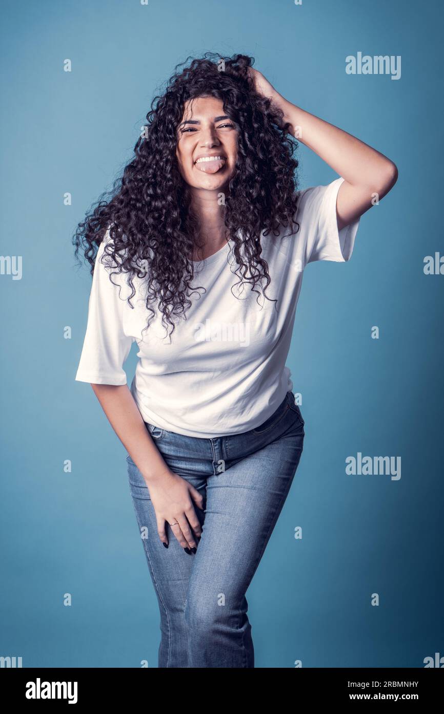 Shot of happy confident curly woman with toothy smile, wears casual basic solid white t-shirt, expresses good emotions, enjoys nice day, making tongue Stock Photo