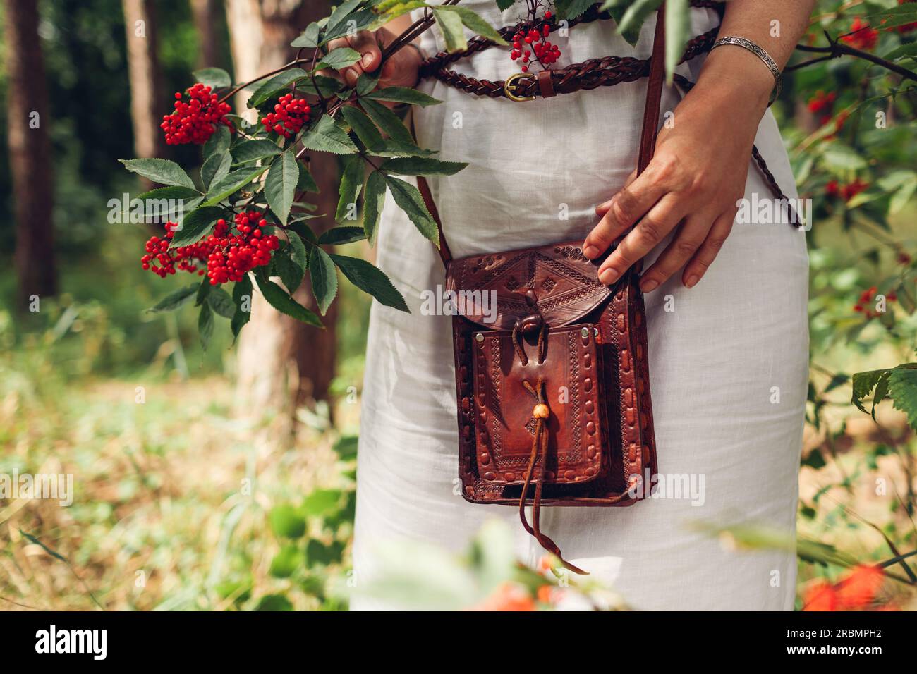 Close up of brown handmade leather purse. Stylish woman wearing white dress, belt, holding rustic handbag by red berries outdoors. Casual boho female Stock Photo