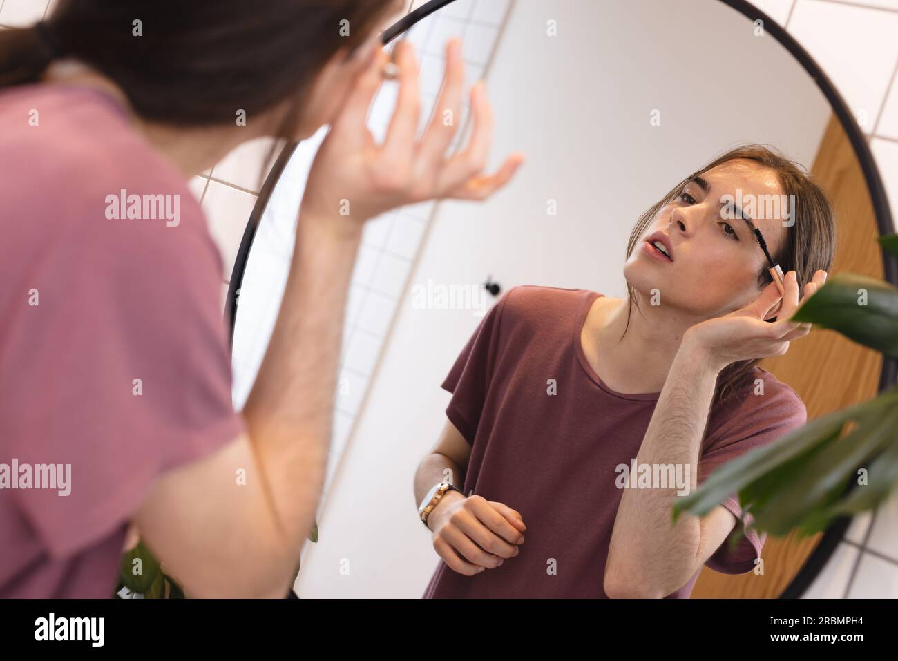 Non-binary transgender woman applying eyebrow makeup looking in bathroom mirror at home. Gender fluidity, health, beauty and lifestyle. Stock Photo