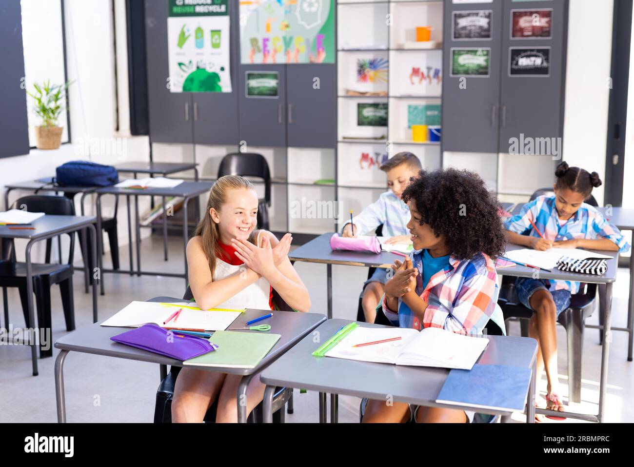 Two smiling, diverse schoolgirls practicing sign language in class ...