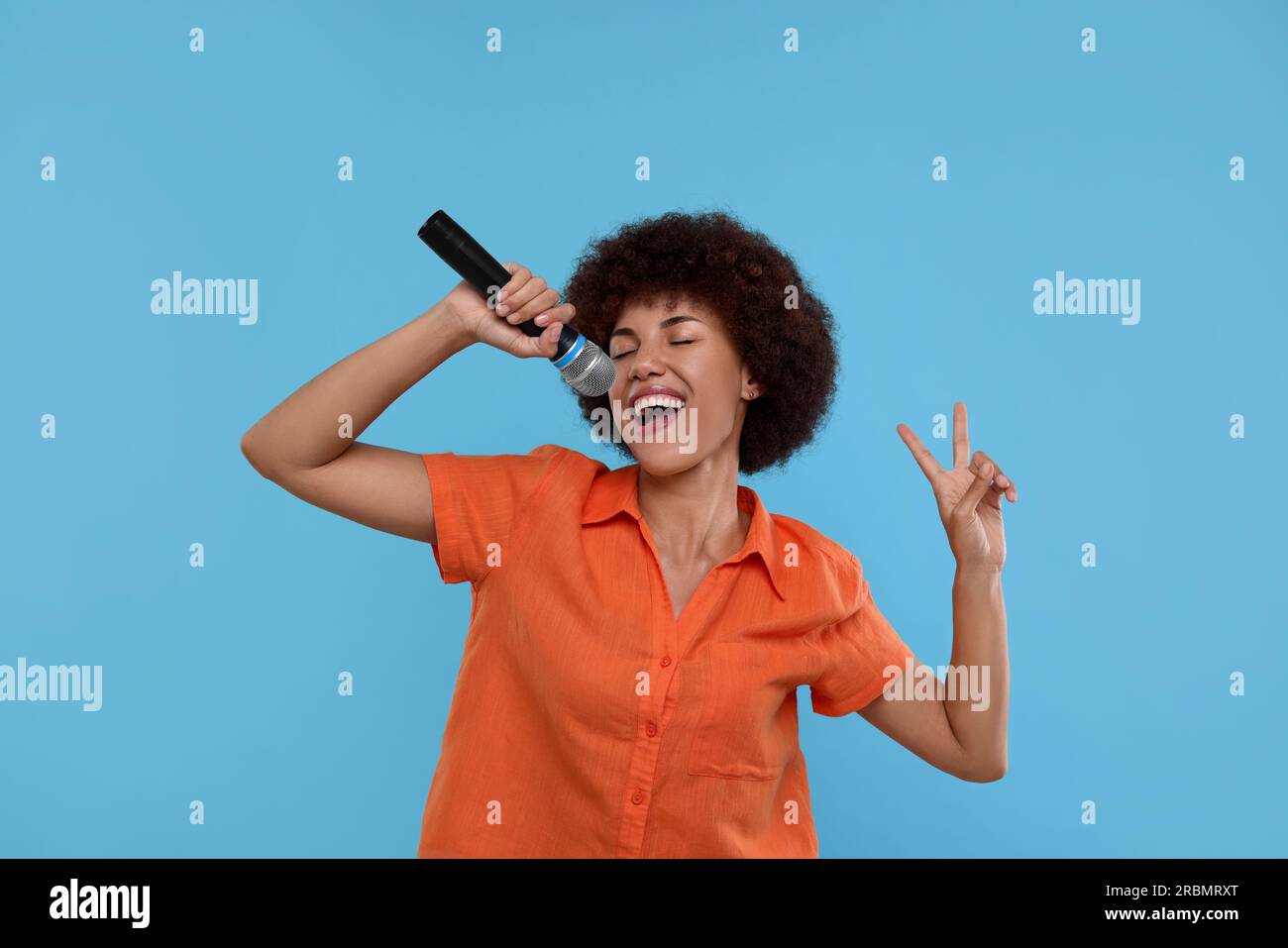 Curly young woman with microphone singing and showing victory sign on light blue background Stock Photo