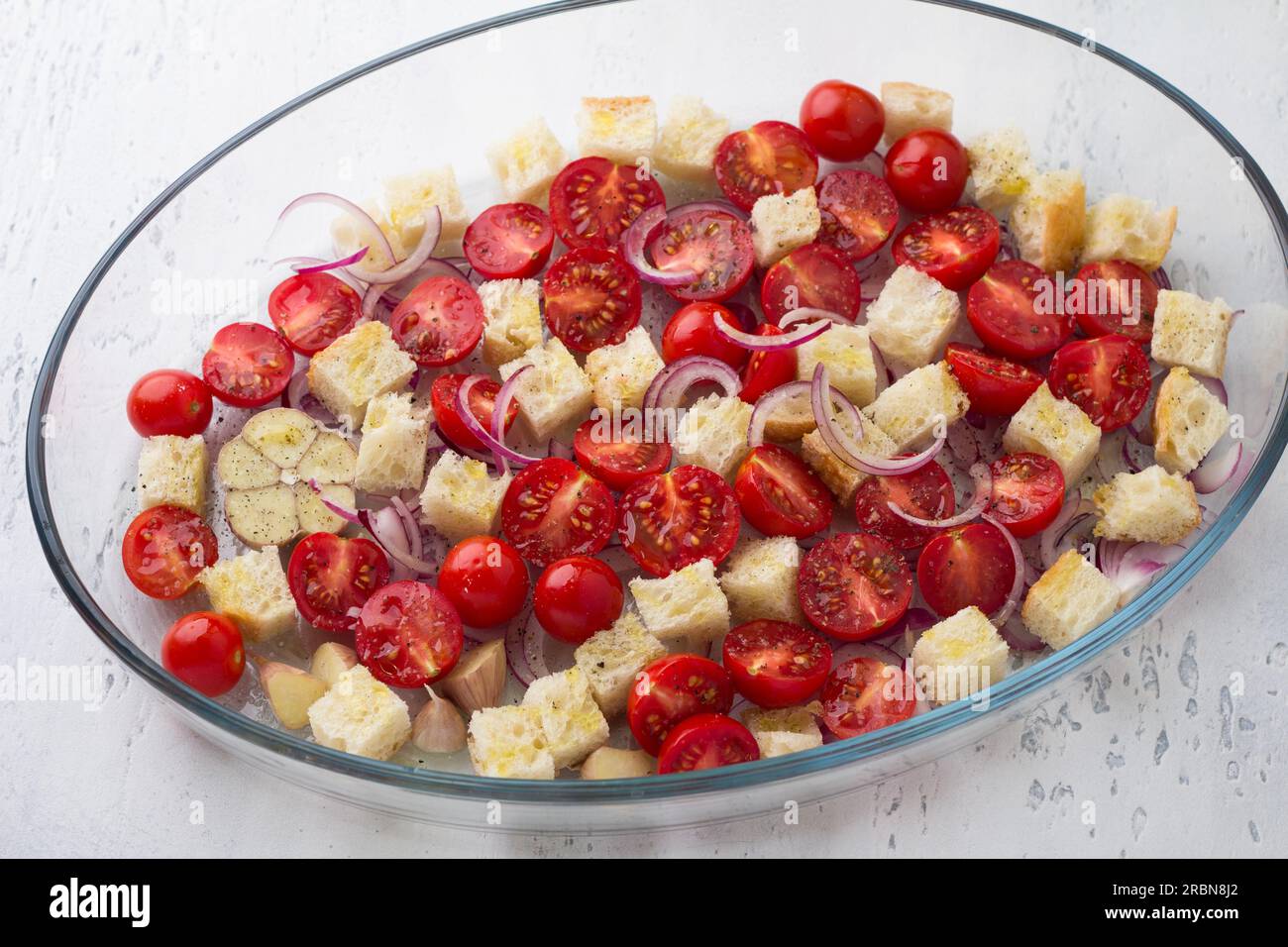 Cherry tomatoes, croutons, red onion, garlic and spices in a glass baking dish on a light gray background, top view. cooking italian panzanella salad Stock Photo