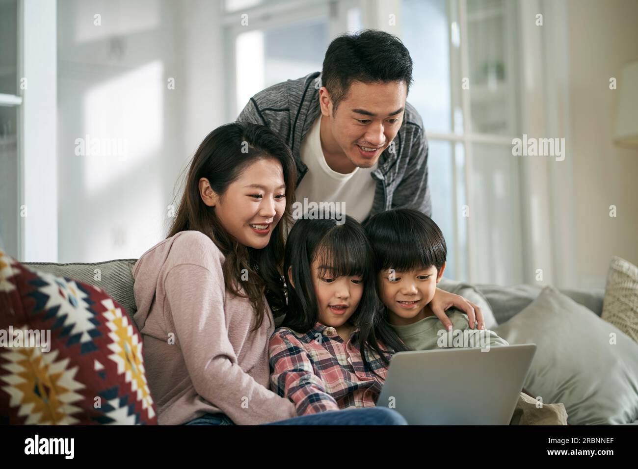 happy young asian couple with two children sitting on family couch at home using laptop computer together Stock Photo