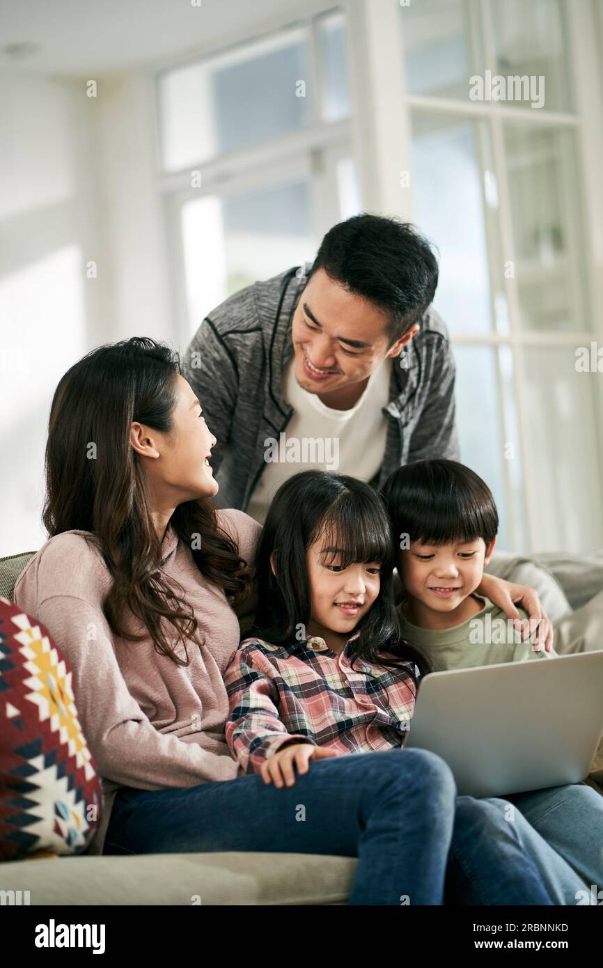 young asian couple and two children sitting on family couch at home watching movie together using laptop computer Stock Photo