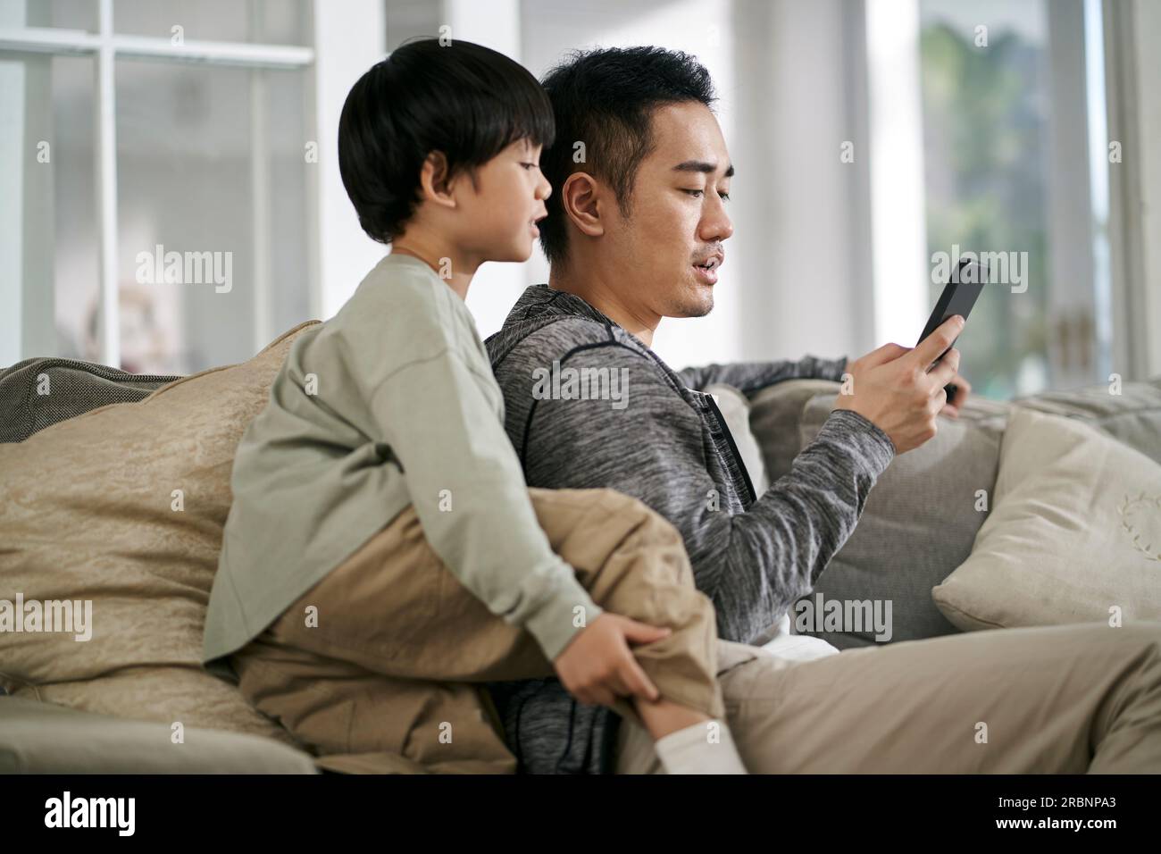 young asian father and son sitting on family couch at home looking at mobile phone together Stock Photo