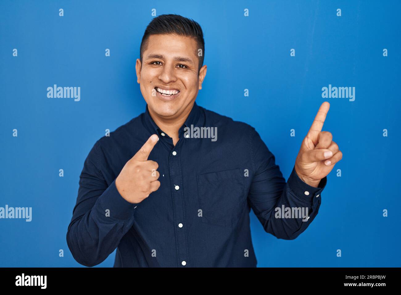 Hispanic young man standing over blue background smiling and looking at ...