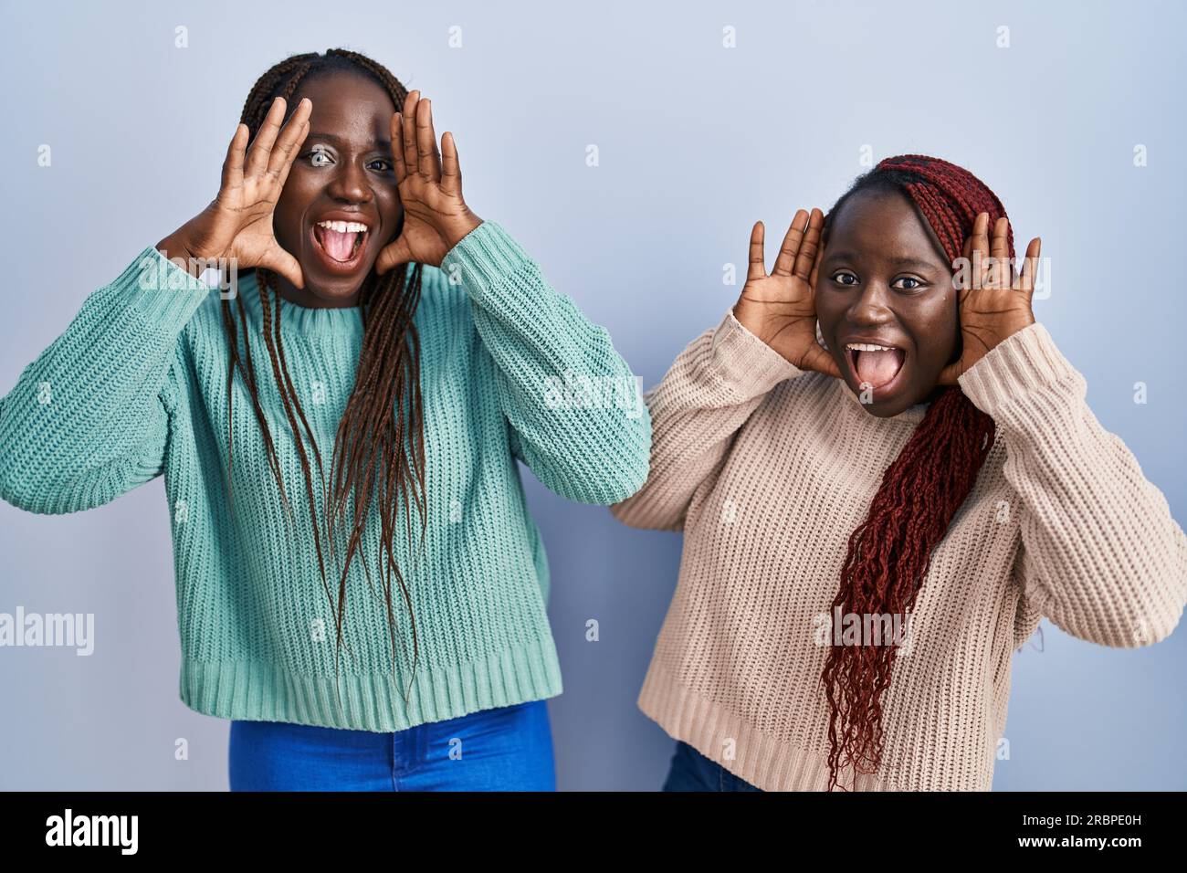 Two african woman standing over blue background smiling cheerful playing peek a boo with hands showing face. surprised and exited Stock Photo