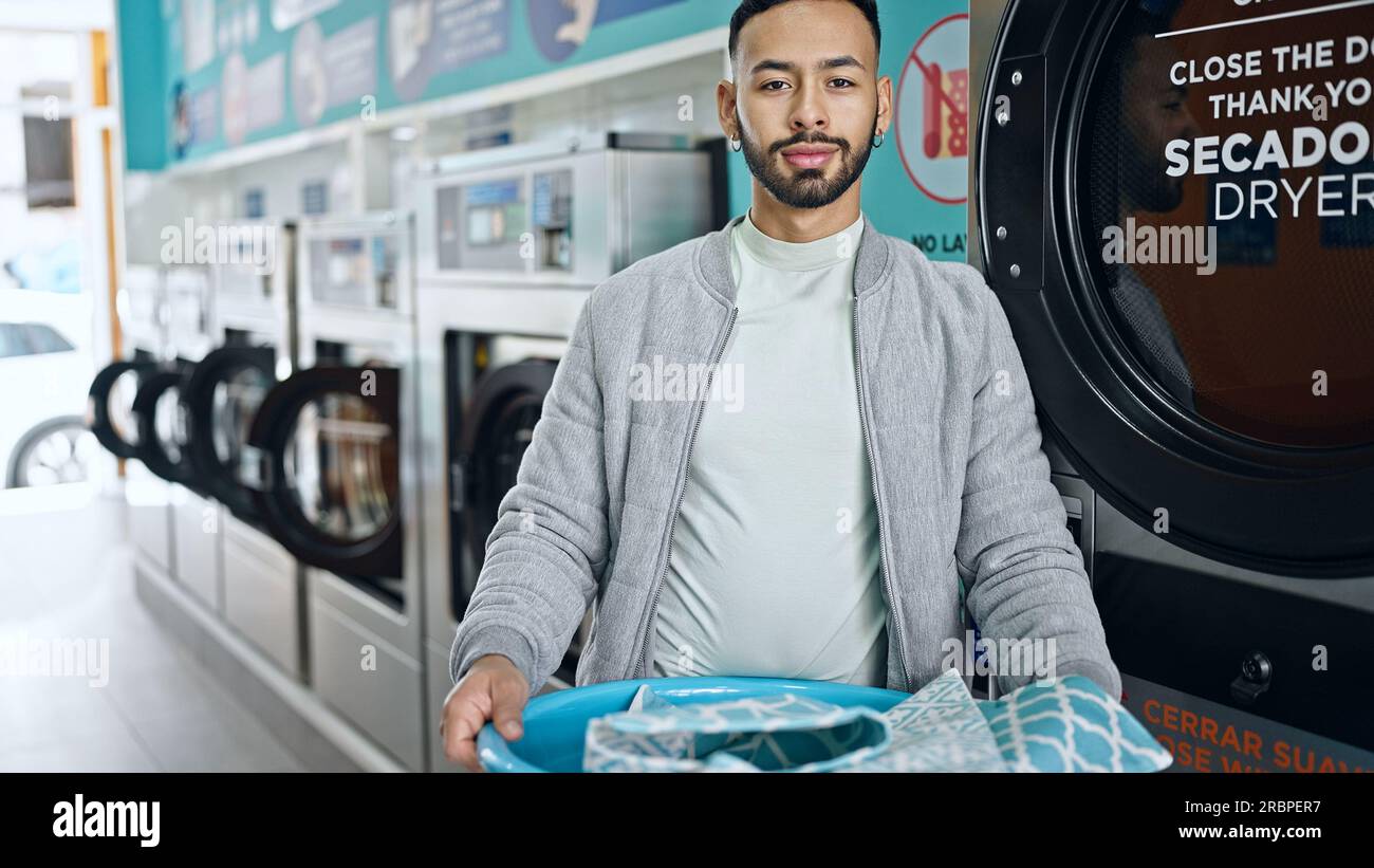 Young hispanic man holding basket with clothes at laundry facility Stock Photo