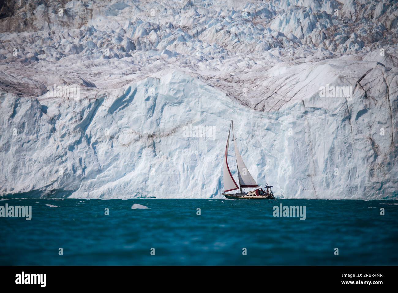 Extreme sail along the face of a massive glacier in East Greenland. Sailboat is sailing among icebergs with a full sail out. Stock Photo