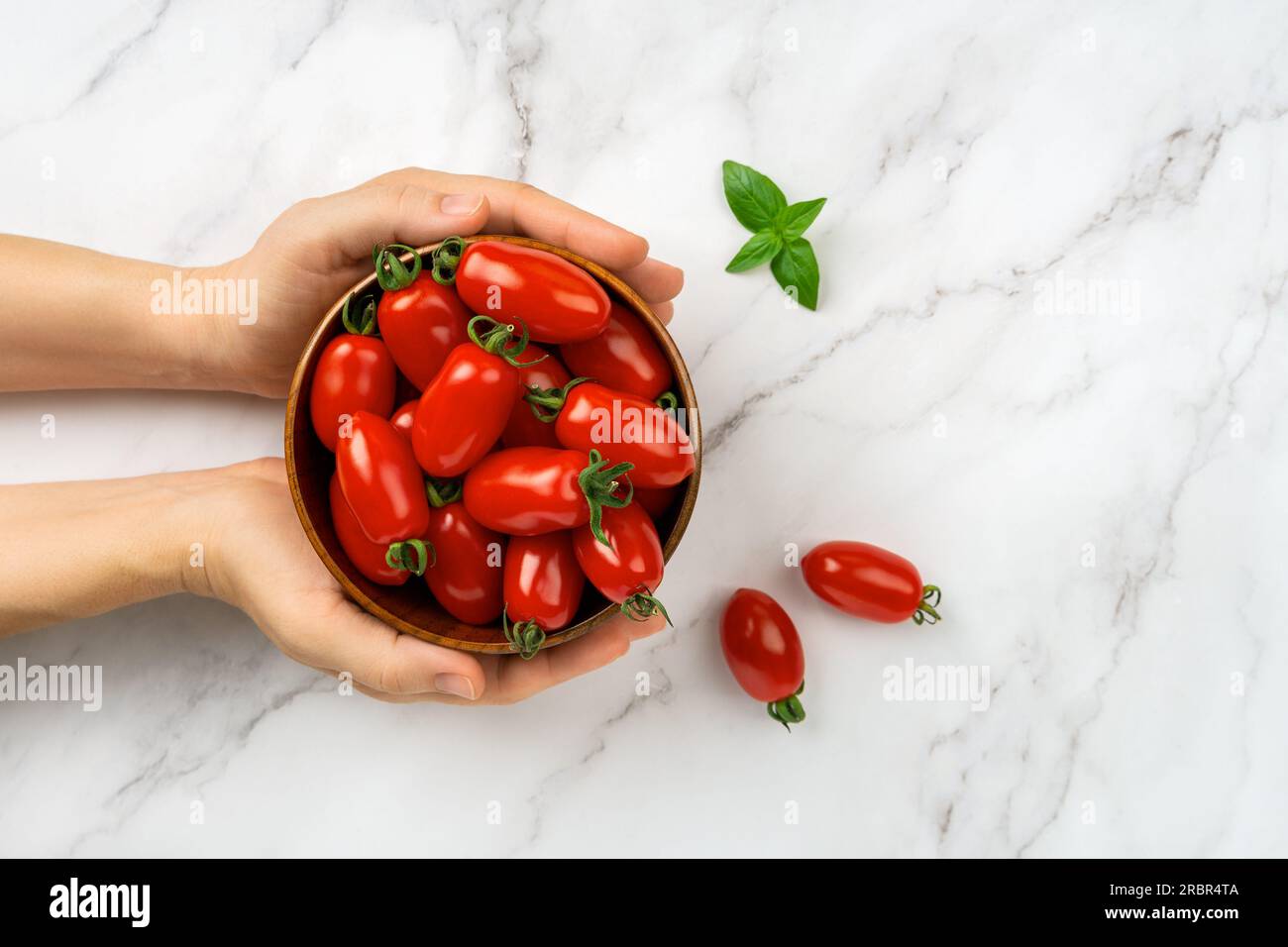 Bowl of fresh Ornela cherry tomatoes in a woman hands over marble background. Female hands hold bowl of red ripe small tomatoes. Organic vegetables. Stock Photo