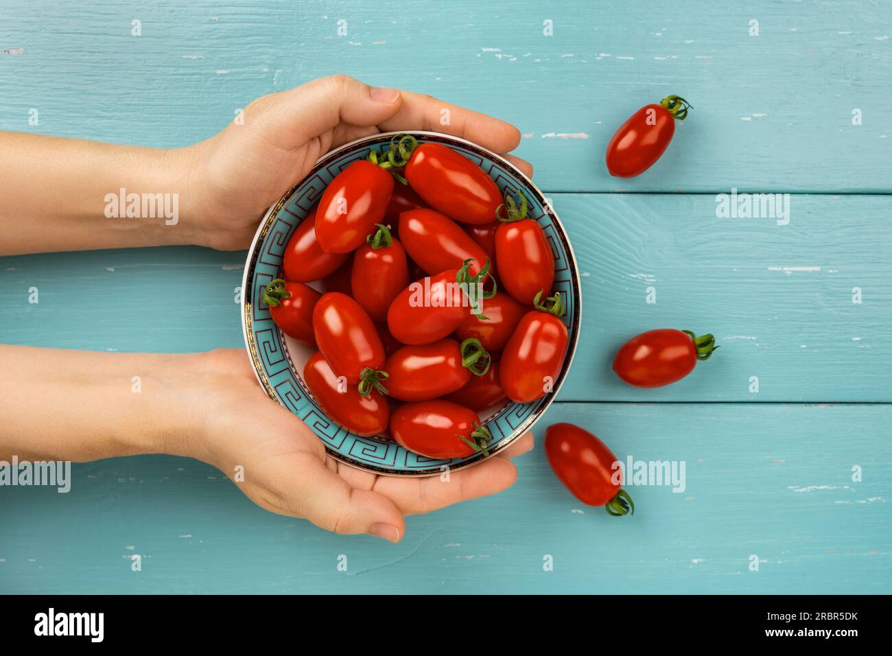 Woman hands hold bowl of red small tomatoes over turquoise wood background. Bowl of ripe Ornela cherry tomatoes in a female hands. Organic vegetables Stock Photo